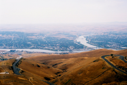 [The lumpy hill is covered in wheat-colored vegeation and no trees. The roadway is a series of switchbacks. On the other side of the hills in the haze are the rivers and buildings and trees.]